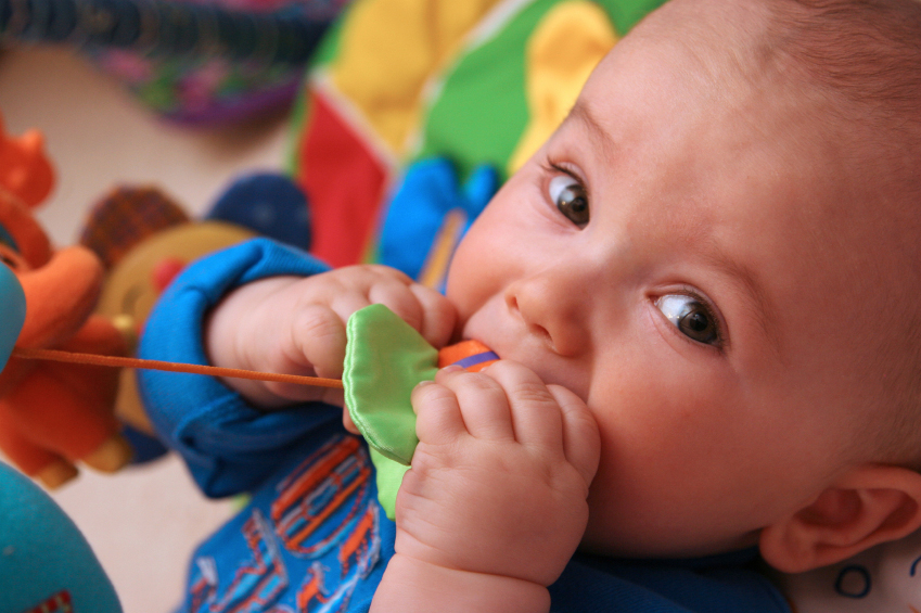 Baby kijkt in camera met kleurrijk speeltje in de handjes en de mond.